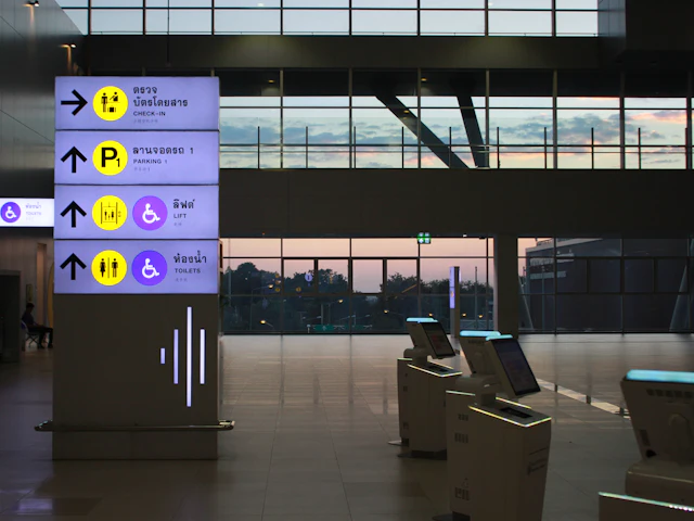A tall lit sign column beside a row of self-service terminals in a hall at dusk.