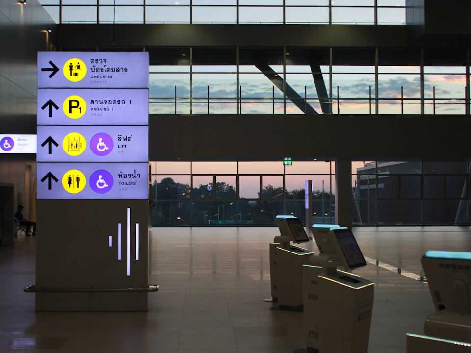 A tall lit sign column beside a row of self-service terminals in a hall at dusk.