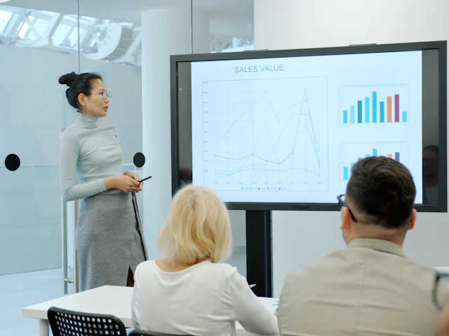 A presenter beside a large screen on a stand, showing charts to two colleagues.